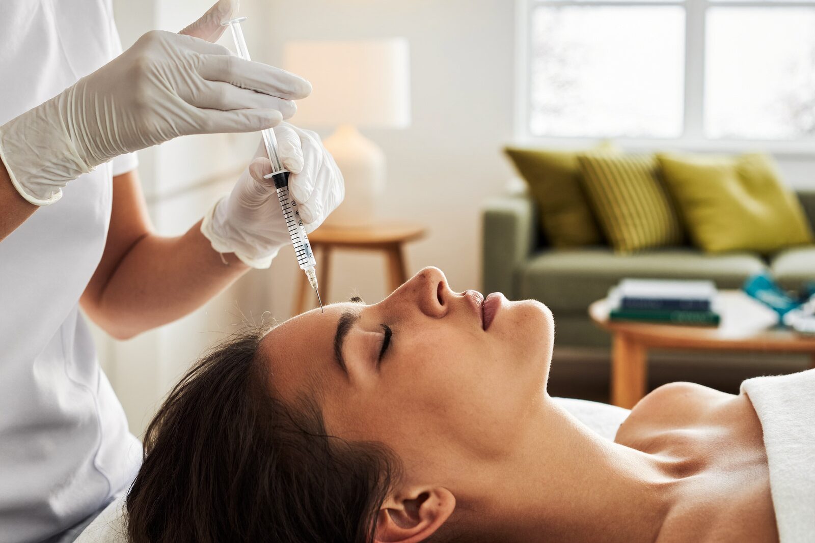 A gloved provider holding a fine syringe above a relaxed client's forehead in a bright living room